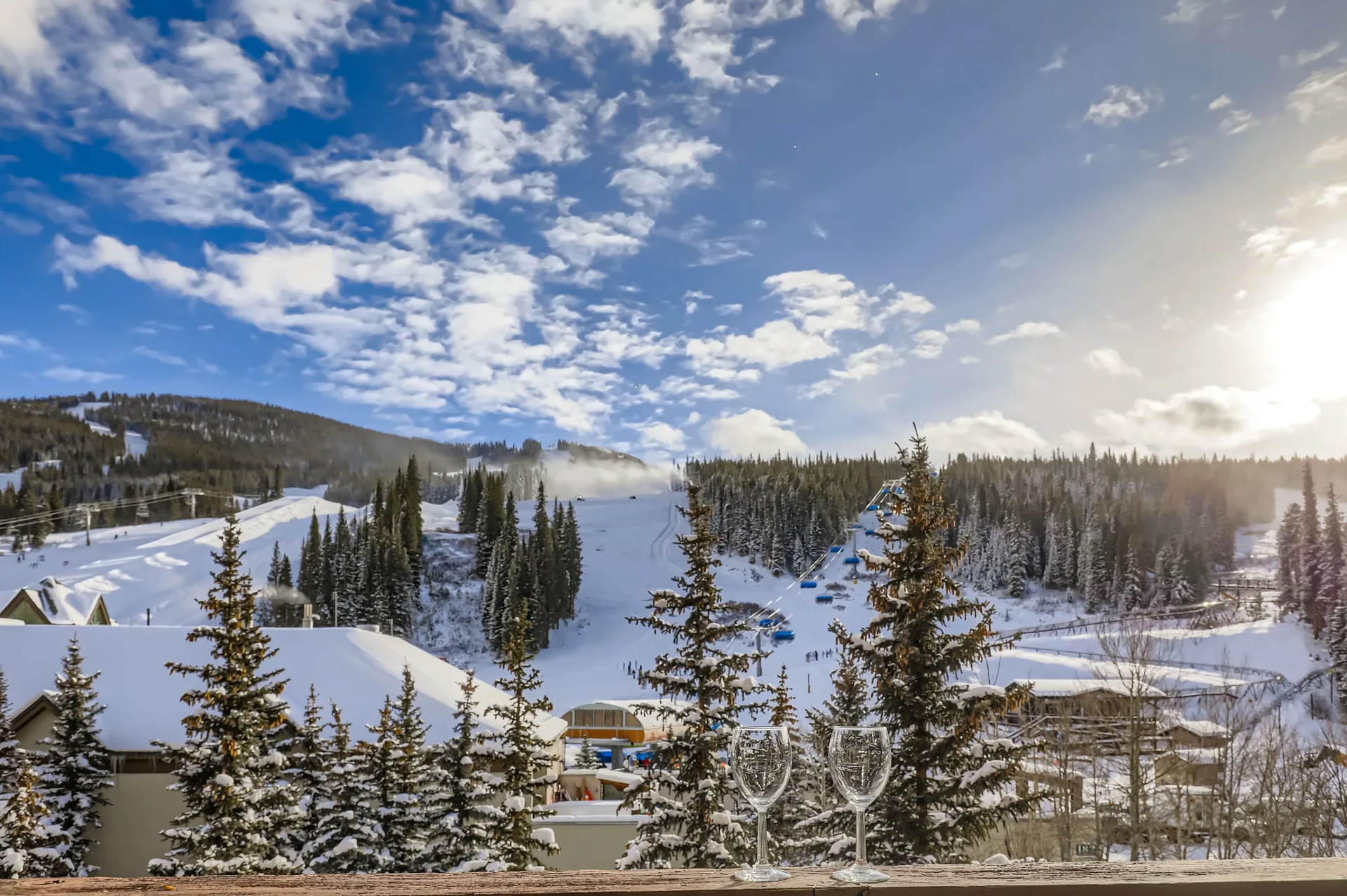 Snowy ski slopes and evergreen trees in morning sunlight, with two wine glasses on a balcony railing